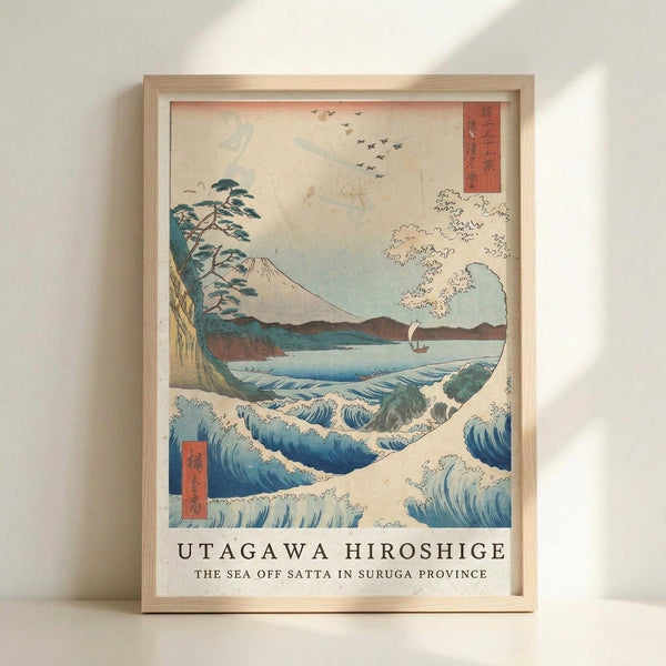 May include: Framed Japanese woodblock print reproduction featuring dramatic ocean waves in blue and white, with Mount Fuji in the background, pine trees on a cliff, small sailing boat, and flying birds. The artwork has traditional ukiyo-e styling with red text cartouches. Displayed in light wood frame against white wall with natural lighting creating soft shadows.