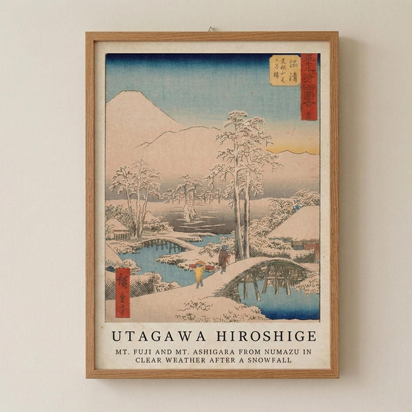 May include: Framed Japanese woodblock print reproduction hanging on cream wall. Shows snowy winter landscape with Mount Fuji in background, snow-covered trees, blue river winding through scene, and small figures crossing wooden bridge. Artwork features soft blues, whites, and earth tones in traditional ukiyo-e style with red text cartouches. Natural wood frame with white matting. Text below image reads "UTAGAWA HIROSHIGE" and describes the scene.