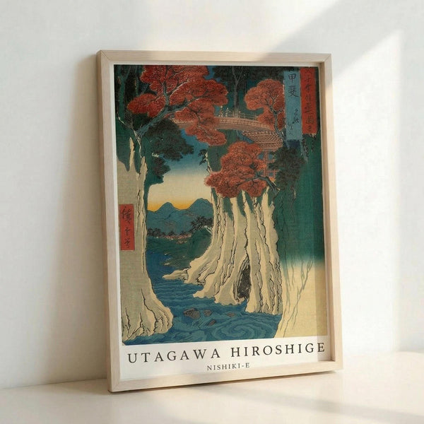 May include: Framed Japanese woodblock print featuring dramatic waterfall cascading down white cliffs with red autumn maple trees above, traditional pagoda structure visible among foliage, mountains in distant background under gradient sky, displayed in light wooden frame against white wall with soft natural lighting