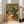 May include: Framed Van Gogh-style poster featuring thick impasto brushstrokes depicting gnarled olive trees with golden-green foliage along a winding dirt path, with a small house visible in the background. The artwork shows swirling, textured paint in earthy yellows, greens, and browns. The print is displayed in a light wood frame leaning against a white wall next to a beige upholstered armchair in a bright, modern living room with large windows.