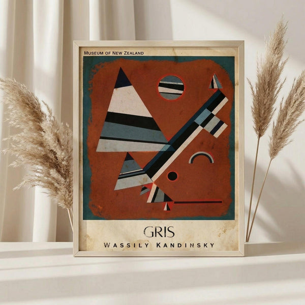 May include: Framed museum poster print of Wassily Kandinsky's abstract artwork featuring geometric shapes in rust orange, black, white, and teal colors. The vintage-style print shows triangular and linear forms with small circles against a weathered background. Displayed on white surface with dried pampas grass stems on both sides and soft beige curtains behind.