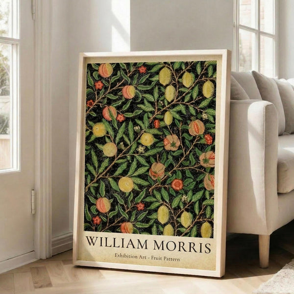 May include: Framed botanical art print leaning against white wall beside beige upholstered chair. Print features dense pattern of green foliage with yellow and orange fruits on dark background, displayed in light wooden frame. Sunlight streams through tall windows onto hardwood floors in bright, airy living room setting.