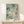 May include: Framed abstract art print with blue-green watercolor background, floating circles and dots in various sizes, vertical dripping effects, and text reading "YAYOI KUSAMA" and "KYOTO" at bottom. Light wood frame displayed on white wooden console table with small white wrapped gift box beside it against cream wall.
