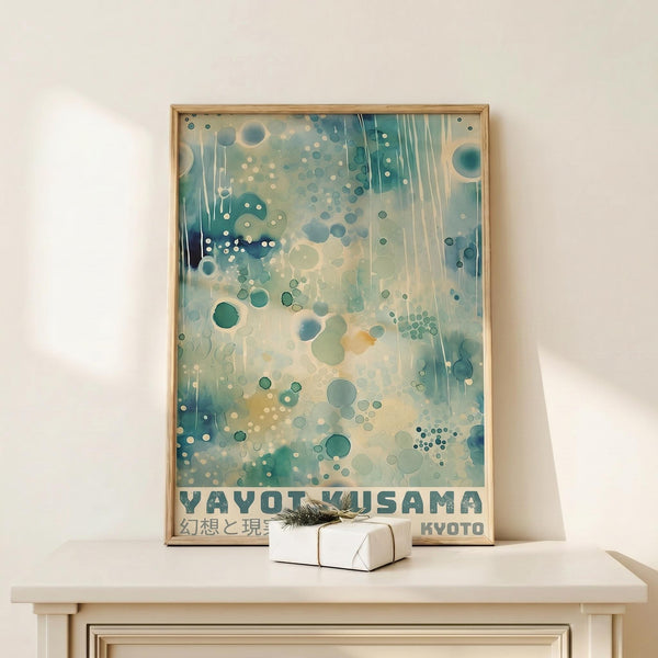 May include: Framed abstract art print with blue-green watercolor background, floating circles and dots in various sizes, vertical dripping effects, and text reading "YAYOI KUSAMA" and "KYOTO" at bottom. Light wood frame displayed on white wooden console table with small white wrapped gift box beside it against cream wall.