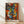 May include: Framed abstract cubist-style artwork featuring geometric faces in vibrant colors including blues, oranges, yellows, and reds, with multiple eyes and fragmented facial features. The wooden-framed poster leans against a white wall on light wooden flooring, with natural sunlight creating soft shadows across the scene.