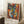 May include: Framed abstract cubist-style artwork featuring geometric faces in vibrant blues, oranges, yellows and reds, displayed in light wooden frame against white wall. Coffee cup on wooden surface and beige fabric visible nearby, creating cozy interior room setting with natural lighting casting shadows.