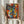 May include: Person in beige ruffled sleeves holding framed abstract cubist-style artwork featuring colorful geometric faces with multiple eyes and fragmented features in blues, reds, yellows, and oranges. Dried wheat stems visible beside the frame against soft white curtained background.