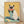 May include: Framed artwork leaning against white wall on wooden floor, showing a cream and dark brown Siamese cat wearing coral sunglasses and holding a watermelon slice. The cat sits on a mint green surface with lemon slices nearby, painted in warm yellows and soft pastels with visible brushstroke texture in a light wood frame.