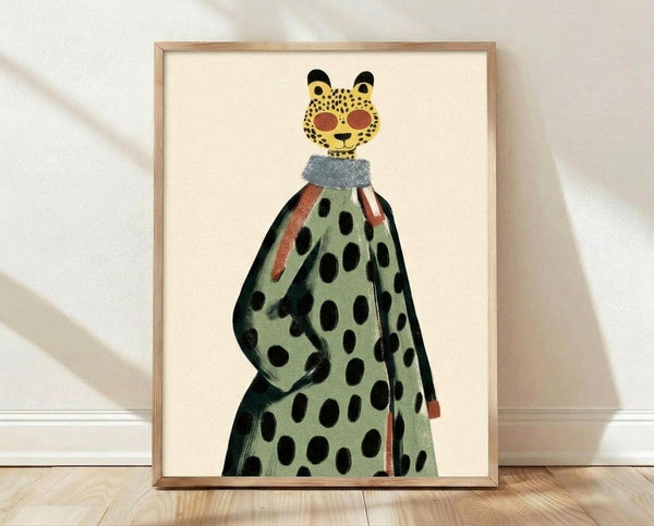 May include: Framed art print leaning against white textured wall on light wood floor. The poster features a whimsical illustration of a leopard head with yellow spotted face and black ears, wearing a blue-gray collar and flowing green cape or dress decorated with large black polka dots and coral-colored stripes. The artwork has a cream background in a thin natural wood frame.