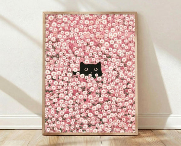 May include: Framed poster print leaning against white wall on light wood floor. Light wood frame contains artwork showing small black cat face with bright eyes peeking through dense field of pink and white daisy flowers. Soft natural lighting creates gentle shadows on textured white wall behind frame.