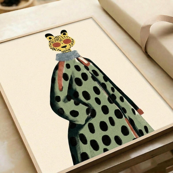 Framed artwork showing a stylized figure wearing a green coat with black polka dots and a leopard face mask. The illustration uses muted colors including sage green, black spots, and coral accents around the collar. The piece is displayed in a light wood frame with white matting, positioned on white paper rolls in soft natural lighting.