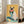May include: Framed artwork leaning against white wall next to beige sofa showing a Siamese cat wearing coral sunglasses and holding a watermelon slice. The painting features a yellow background with teal and blue areas, and halved limes at the bottom. The cat has black ears, face markings, and paws with a cream-colored body. Natural lighting from nearby windows illuminates the bright, playful artwork in this modern living room setting.