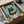 May include: Framed art print showing a black cat with yellow eyes sitting on a pink toilet while reading a green book. The illustration features a teal tiled bathroom background with potted plants in terracotta pots. The artwork is displayed in a black frame, packed in a cardboard shipping box with protective wrapping materials. The whimsical cartoon-style print uses a limited color palette of mint green, coral pink, and black.