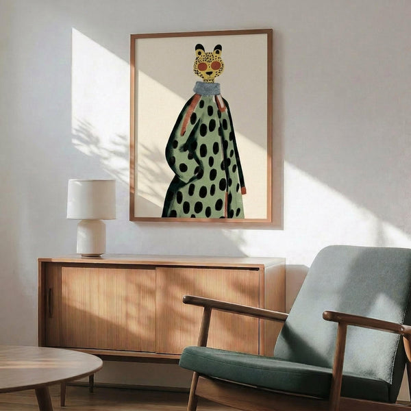 May include: Framed art print displaying a stylized leopard figure wearing a polka-dotted green dress or coat with blue collar detail, mounted on white wall above wooden sideboard. Mid-century modern room setting features sage green upholstered armchair with wood frame, round side table, white ceramic table lamp, and natural sunlight casting geometric shadows across the scene.