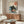 May include: Japanese-style art print displayed above wooden credenza in modern living room. Black cat with yellow eyes eating ramen from bowl, surrounded by blue ocean waves in traditional ukiyo-e style. Red sun and Japanese characters in upper corner. Print appears framed and hung on white wall. Glass coffee table reflects the artwork's wave pattern. White curved chair and brass floor lamp visible in scene.