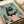 Illustrated print featuring a black cat with yellow eyes reading a green book while sitting on a pink toilet. The scene shows a bathroom with mint green tile walls and terracotta planters containing various plants like cacti and ferns. The artwork has a textured, vintage print style with muted colors and appears to be displayed flat against white paper with a beige ribbon visible at the edge.