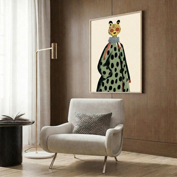 May include: Whimsical illustration of a leopard head with yellow spotted fur and black ears atop a human figure wearing a sage green coat with black polka dots and coral-colored sleeves. The artwork is displayed in a light wood frame on wood-paneled walls above a cream upholstered armchair with geometric patterned pillow, brass floor lamp, and dark round side table with open book.