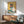May include: Framed poster artwork showing a white and dark Siamese cat wearing orange sunglasses, holding a watermelon slice, painted in watercolor style with yellow background and blue-green surface. Wood-framed print displayed above mid-century modern walnut credenza with white ceramic lamp, next to sage green upholstered armchair in sunlit room with white walls.