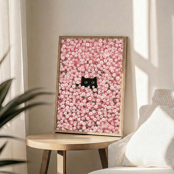 May include: Framed poster displaying a pink and white daisy flower pattern with a small black cat peeking through the blooms. The artwork is shown in a thin gold frame, positioned on a round wooden side table next to a beige upholstered chair. Soft natural lighting creates gentle shadows across the neutral-toned room setting with white walls and a plant visible in the background.