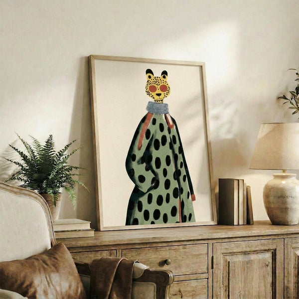 May include: Framed art print displaying a whimsical illustration of a leopard head with yellow spotted face and black ears, wearing a flowing green dress covered in black polka dots with coral-colored accents. The artwork sits on a rustic wooden console table alongside books, a ceramic vase, and fern plant, with a beige upholstered chair visible in the foreground of this neutral-toned living room setting.
