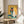 May include: Framed artwork showing a white and black Siamese cat wearing orange sunglasses and holding a watermelon slice, painted in vibrant colors against a yellow background with lime slices nearby. The piece sits on a round wooden side table next to a beige upholstered chair and plant in a bright, minimalist room with white walls and natural sunlight streaming in.