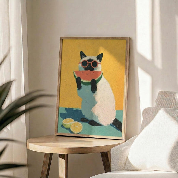 May include: Framed artwork showing a white and black Siamese cat wearing orange sunglasses and holding a watermelon slice, painted in vibrant colors against a yellow background with lime slices nearby. The piece sits on a round wooden side table next to a beige upholstered chair and plant in a bright, minimalist room with white walls and natural sunlight streaming in.