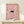 May include: Framed poster print showing a black cat with bright eyes peeking through a dense field of pink and white daisy flowers. The artwork has a whimsical, dreamy quality with soft pink tones throughout. Displayed in a light wood frame on a cream-colored wooden shelf against a white wall, with a small wrapped gift box beside it.