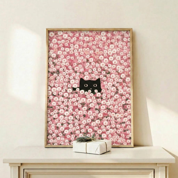 May include: Framed poster print showing a black cat with bright eyes peeking through a dense field of pink and white daisy flowers. The artwork has a whimsical, dreamy quality with soft pink tones throughout. Displayed in a light wood frame on a cream-colored wooden shelf against a white wall, with a small wrapped gift box beside it.