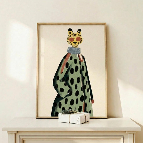 May include: Framed art print showing a whimsical leopard character wearing a flowing green polka dot dress or cape with orange accents and a gray collar. The leopard has a yellow spotted face with orange cheeks and black ears. The artwork is displayed in a thin wooden frame on a light wood shelf against a cream wall, with a small wrapped gift box positioned in front.