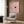 May include: Modern living room with beige armchair and geometric pillow, brass floor lamp, dark round side table with open book, wood paneled wall, and framed poster showing pink cherry blossoms with small black cat face peeking through the flowers in light wood frame.