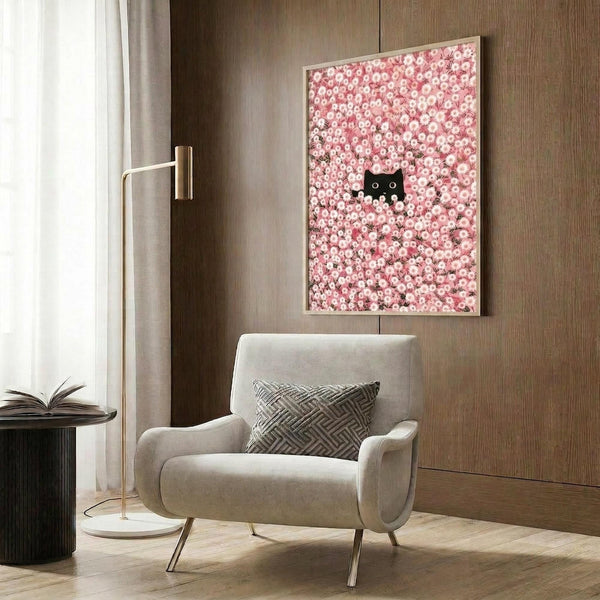 May include: Modern living room with beige armchair and geometric pillow, brass floor lamp, dark round side table with open book, wood paneled wall, and framed poster showing pink cherry blossoms with small black cat face peeking through the flowers in light wood frame.