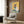 May include: Framed poster artwork displayed on wood paneled wall above beige upholstered armchair. The painting shows a Siamese cat wearing round sunglasses holding a watermelon slice, with lemon halves nearby, rendered in warm yellow, teal, and coral tones. Modern living room setting with floor lamp, side table with open book, and geometric patterned throw pillow.