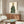 May include: Contemporary living room with wooden sideboard displaying framed artwork of stylized leopard figure wearing green polka dot dress with blue collar. White upholstered chair, brass floor lamp, clear table lamp, and oval glass coffee table with colorful abstract art visible underneath complete the mid-century modern interior scene.