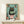 May include: Framed poster print showing a black cat with yellow eyes reading a green book while sitting on a pink toilet, surrounded by potted plants including cacti and succulents against a turquoise tiled wall background. The artwork has a whimsical illustration style with textured details. Frame appears in light wood finish, displayed on a light wooden shelf with a small wrapped gift box beside it.