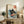 May include: Framed art print featuring a black cat with yellow eyes eating ramen from a white bowl with red garnishes, surrounded by stylized blue ocean waves in traditional Japanese ukiyo-e style. The print has Japanese characters in a red circle and cream background. Displayed on a rustic wooden sideboard next to books, a potted fern, ceramic vase, with a beige armchair visible in a warmly lit living room setting.