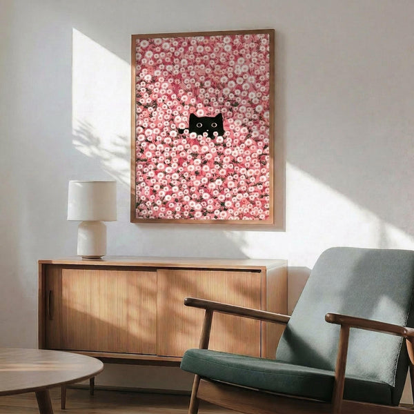 May include: Framed artwork showing a black cat with green eyes peeking through a dense field of pink cherry blossoms or daisies, displayed above a mid-century modern wooden credenza with white ceramic lamp. Green upholstered armchair with wooden arms sits nearby in minimalist living room with white walls and natural sunlight streaming across the space.