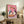 May include: Framed artwork displaying two white geese wearing colorful scarves and sunglasses sharing pizza and wine on a pink background, positioned on a rustic wooden sideboard alongside books, potted plants, and ceramic vases in a cozy living room setting with neutral tones and natural lighting.