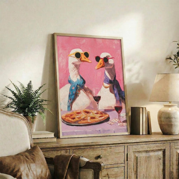 May include: Framed artwork displaying two white geese wearing colorful scarves and sunglasses sharing pizza and wine on a pink background, positioned on a rustic wooden sideboard alongside books, potted plants, and ceramic vases in a cozy living room setting with neutral tones and natural lighting.