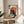 May include: Framed poster featuring a black cat with white eyes eating ramen noodles with chopsticks, set against an orange sun backdrop in traditional Japanese art style. The artwork displays Japanese text and shows a bowl of ramen with an egg. The wooden-framed print sits on a light wood round side table next to a beige upholstered chair, with a plant casting shadows on the white wall behind in natural sunlight.