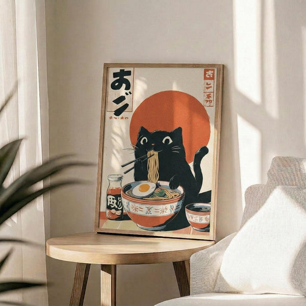 May include: Framed poster featuring a black cat with white eyes eating ramen noodles with chopsticks, set against an orange sun backdrop in traditional Japanese art style. The artwork displays Japanese text and shows a bowl of ramen with an egg. The wooden-framed print sits on a light wood round side table next to a beige upholstered chair, with a plant casting shadows on the white wall behind in natural sunlight.