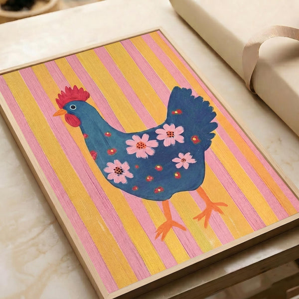 May include: A framed folk art poster featuring a decorative blue hen with red comb and orange feet, adorned with pink flowers and small red dots on its body, displayed against alternating pink and yellow vertical stripes. The artwork is shown in a light wooden frame on a beige sofa next to cream pillows in a neutral-toned living room setting.