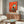 May include: Modern living room mockup showing vibrant orange artwork featuring black and white tuxedo cat sitting on turquoise chair with small birds perched around it. Art displayed above wooden sideboard with sliding doors, accompanied by cream upholstered chair and oval glass coffee table on light wooden floor.