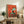 May include: Framed poster artwork displayed on rustic wooden sideboard showing black and white tuxedo cat sitting on turquoise chair with small birds perched above and below, set against vibrant orange-red textured background. Room styled with fern plant, ceramic vase, stacked books, and leather furniture.