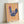 May include: Framed folk art poster featuring a blue hen with red comb and wattle, decorated with pink flowers across its body, standing on orange legs against yellow and pink vertical striped background. The artwork is displayed in a light wood frame leaning against a white wall.