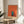 May include: Framed contemporary art print displayed on light wood round side table next to beige upholstered chair. Artwork shows minimalist illustration of person reading in teal doorway against coral orange and cream geometric background. Natural lighting creates soft shadows on white walls, with plant leaves visible at left edge.