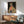 May include: Framed artwork on white wall showing stylized leopard wearing green sunglasses, coral scarf, and spotted coat against dark green background. Modern mid-century living room with sage green upholstered chair, wooden sideboard, small table lamp with white shade. Warm natural lighting creates geometric shadows across the minimalist space.