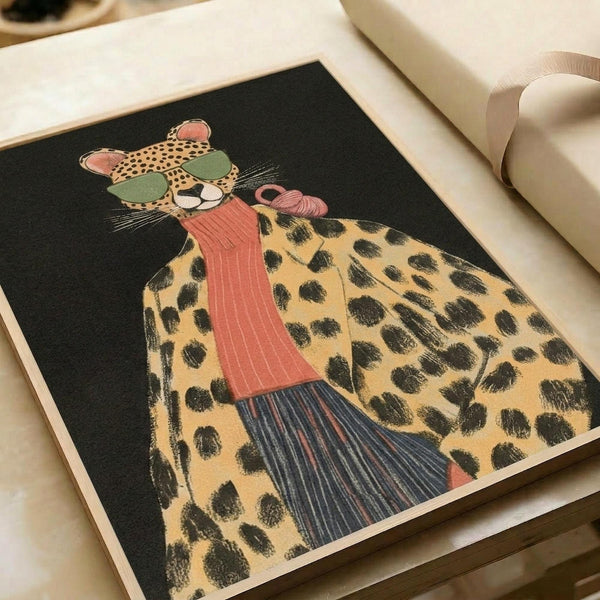 Illustrated cheetah wearing green sunglasses and coral pink scarf on black background. The stylized big cat has yellow fur with black spots and whiskers. Art print displayed in open book or portfolio with cream colored pages and neutral ribbon visible at edges.