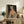 May include: Framed artwork displaying a stylized leopard wearing green sunglasses, coral ribbed turtleneck, and spotted yellow coat against dark background. Print sits on rustic wooden sideboard alongside books, ceramic vase, and potted fern. Cream armchair with leather pillow visible in foreground of neutral-toned living room setting.