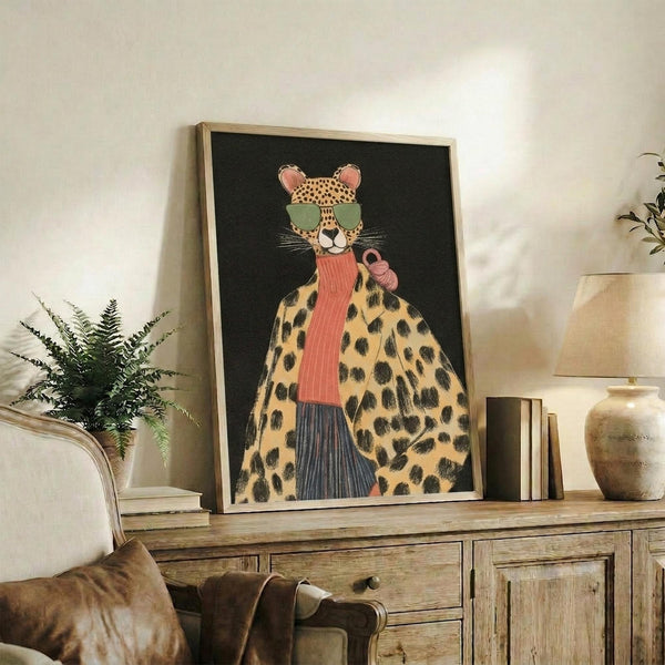May include: Framed artwork displaying a stylized leopard wearing green sunglasses, coral ribbed turtleneck, and spotted yellow coat against dark background. Print sits on rustic wooden sideboard alongside books, ceramic vase, and potted fern. Cream armchair with leather pillow visible in foreground of neutral-toned living room setting.