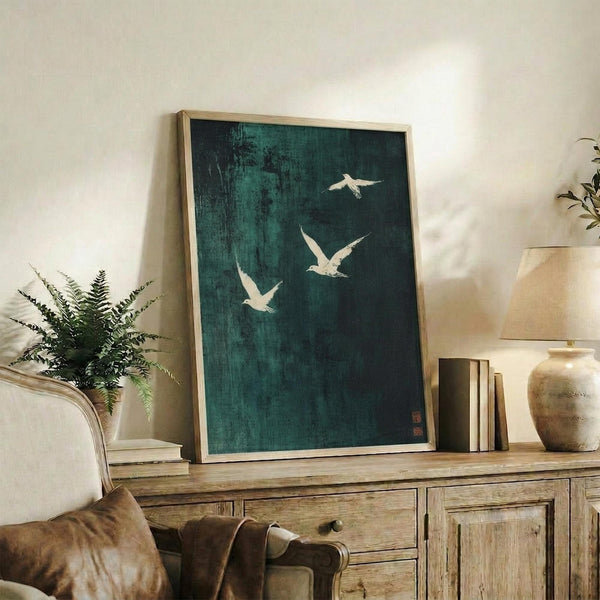 May include: Framed artwork with dark teal background featuring three white birds in flight, displayed on rustic wooden sideboard alongside fern plant, ceramic vase, and stacked books in warm-toned living room setting